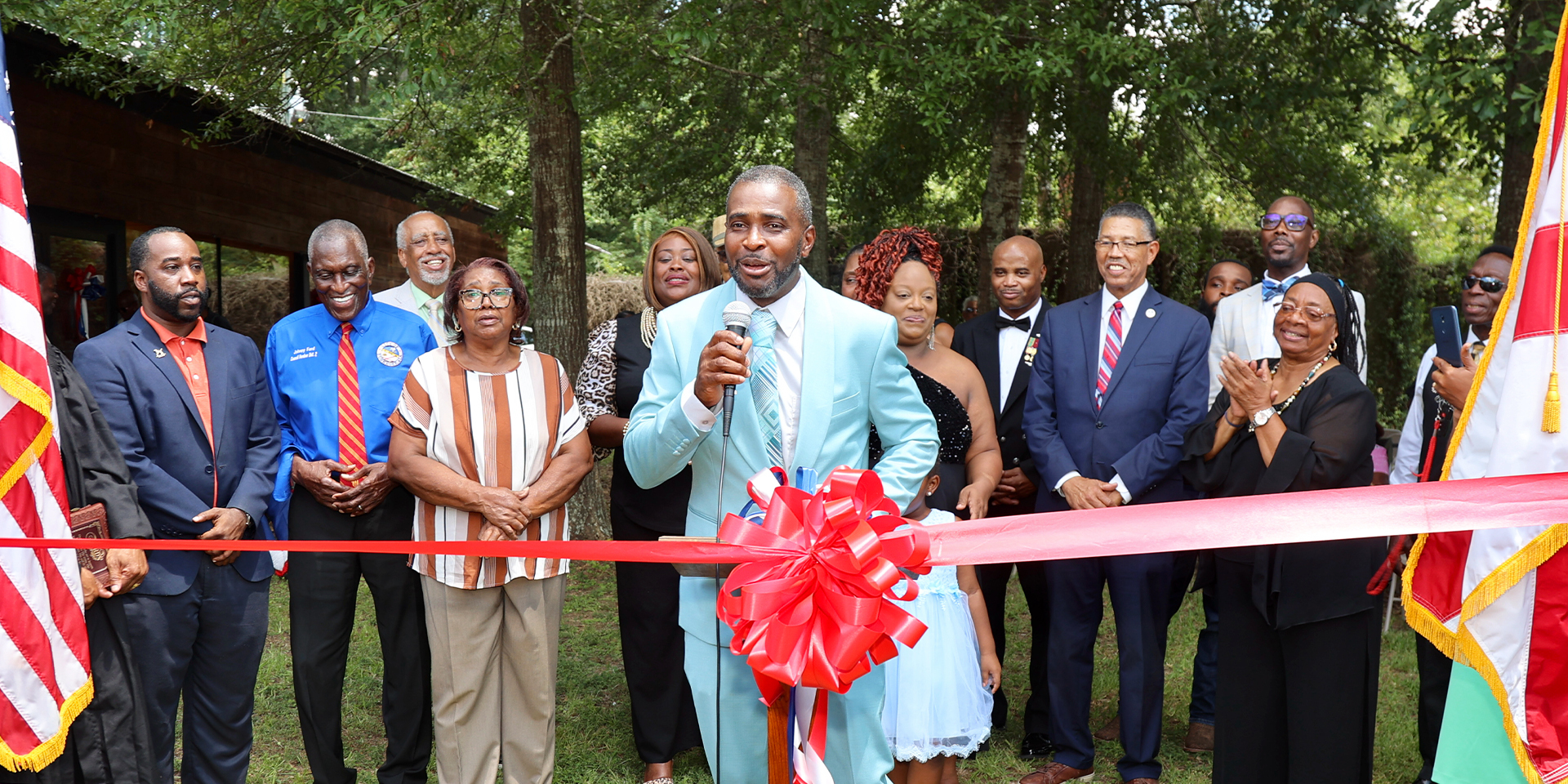 Person in vibrant suit stands in front of crowd behind large ribbon at an outdoor ceremony.